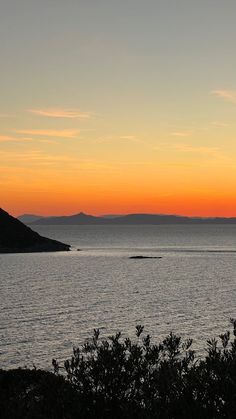 the sun is setting over the ocean with mountains in the distance and trees to the side