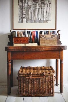 a wooden table topped with books next to a basket under a framed painting on the wall