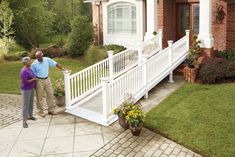 two people standing in front of a house with white railings and brick walkway leading up to the porch