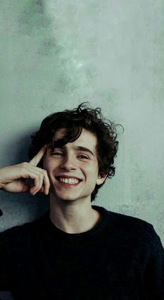 a young man smiling and leaning against a wall with his hand on his head while wearing a black shirt