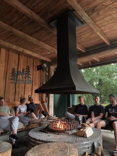 a group of people sitting around a fire place in a wooden building with a metal chimney