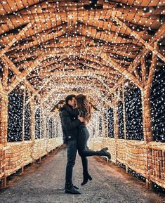 a man and woman standing under a wooden structure covered in fairy lights, with their arms around each other