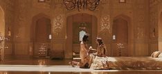 two women sitting on a bed in a large room with chandeliers hanging from the ceiling