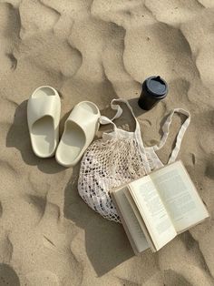 an open book sitting on top of sand next to slippers and a bag filled with books