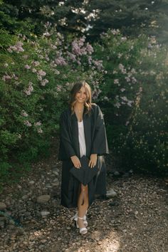 a woman wearing a black robe and white shoes standing in front of some bushes with purple flowers