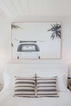 a white bed topped with two pillows next to a painting on top of a wall