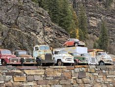 several old trucks are lined up on the side of a mountain road in front of a stone wall