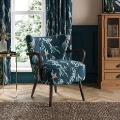 a blue chair sitting in front of a window next to a wooden table and bookcase