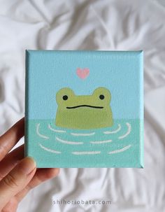 a hand holding up a small square painting with a frog in the water