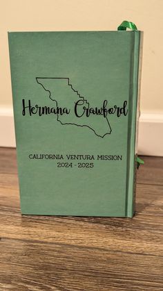 a green book sitting on top of a wooden floor next to a white wall with the words, hermana cranford california ventura mission