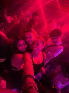 three girls are posing for the camera in front of a crowd at a party with red and pink lights