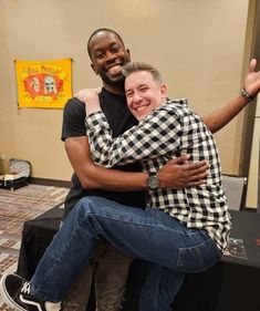 two men hugging each other while sitting on top of a stool in front of a table