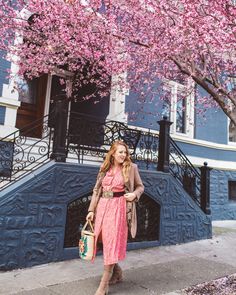 a woman in a pink dress is standing on the sidewalk under a tree with purple flowers