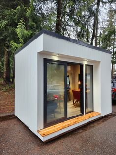 a small white building with sliding glass doors on the front and side, sitting next to a wooded area