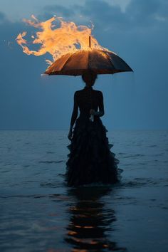 a woman standing in the water holding an umbrella over her head with fire coming out of it