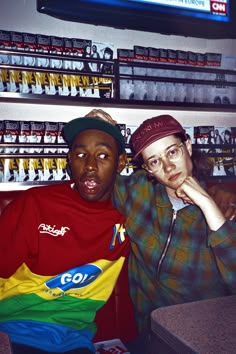 two young men sitting next to each other in front of a shelf with various items on it