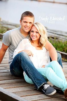 a man and woman are sitting on a wooden dock by the water, smiling at the camera