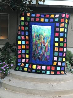 a colorful quilt hanging on the side of a house in front of a flower pot