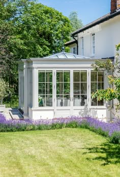 a white house sitting in the middle of a lush green field with purple flowers around it