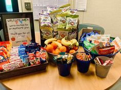 a wooden table topped with buckets filled with fruit and veggies next to a welcome back sign