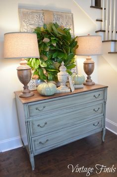 a white dresser with two lamps on top of it next to a stair case and green plant