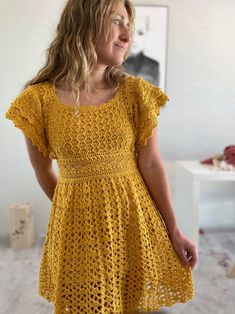 a woman wearing a yellow crochet dress standing in front of a white wall