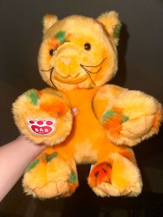 a hand holding a yellow teddy bear with green and orange paint on it