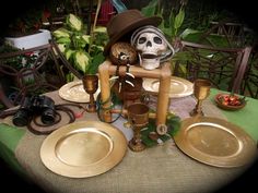 a table topped with lots of gold plates and cups next to a skull statue on top of a wooden stand