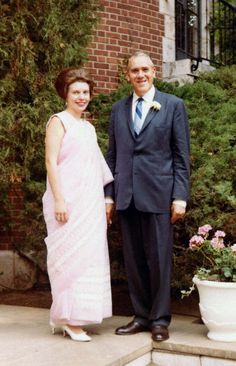 an older man and young woman standing on steps in front of bushes with pink flowers