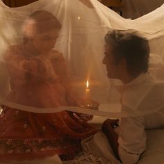 two people sitting on a bed with veils over their heads and candles in the background