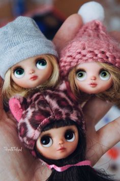 three dolls are posed in the palm of someone's hand, wearing knitted hats and scarves