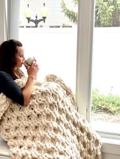 a woman sitting on a window sill wrapped in a chunky blanket and drinking coffee