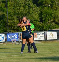 two female soccer players hugging each other in the middle of a field with trees in the background