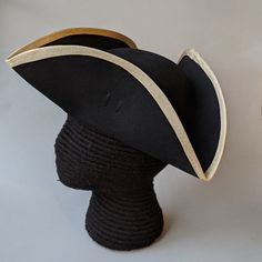 "This military style three cornered tricorn hat is made from top quality wool felt. The edges have been trimmed even and the sides held up or \"cocked\". It has been trimmed around the brim with gold fold-over braid. It is also available with black or white braid. Plain untrimmed hats are listed separately. Gold tone of edging may vary due to availability. Attached is a 3.5 \" black, pleated cockade with gold star button. The brim measures about 4.25\" to 4.5\" and the crown is about 4\" high. W Fitted Black Hat With Structured Crown, Black Fitted Costume Hat With Curved Brim, Classic Black Hat For Costume, Black Brimmed Hat For Ceremonial Occasions, Black Curved Brim Costume Hat, Fitted Costume Hats With Curved Brim, Fitted Brimmed Costume Hats, Black Brimmed Hat For Costume, Fitted Costume Hats And Headpieces With Curved Brim