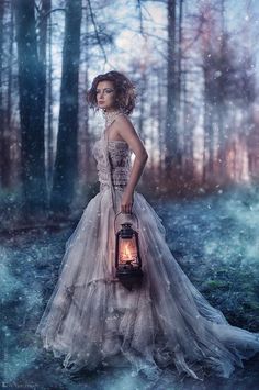 a woman in a dress holding a lantern standing in the woods with snow falling on her