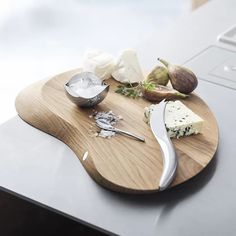 a wooden cutting board topped with cheese next to a knife and bowl of fruit on top of a table