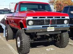 a red truck parked in a parking lot next to other cars and trucks with large tires