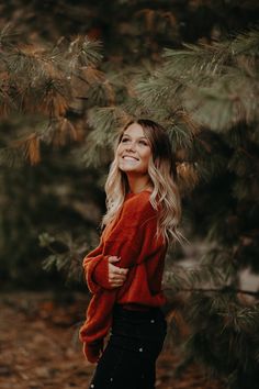 a woman standing in front of pine trees with her arms crossed and smiling at the camera