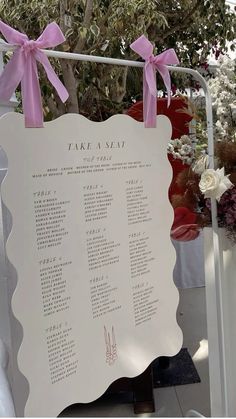 a white sign with pink bows on it that says take a seat in front of some flowers