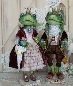 two frog dolls dressed up as princess and frog prince standing next to each other with flowers in their hands