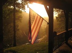 an american flag hanging from the side of a wooden porch with sun shining through trees