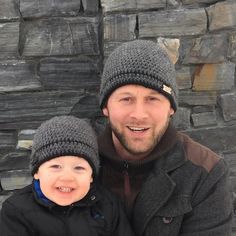 a man holding a small child in front of a stone wall wearing knitted hats