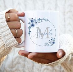 a person holding a white coffee mug with the letter m on it and blue flowers