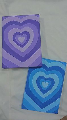 two heart shaped paintings sitting on top of a white bed sheet next to each other