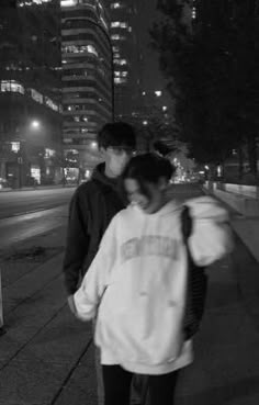 two people are walking down the sidewalk in front of some tall buildings at night time