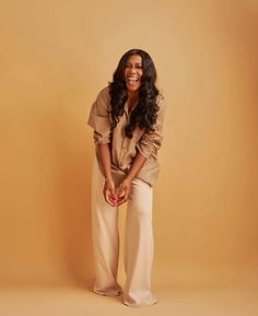 a woman standing in front of a tan background wearing wide legged pants and a brown shirt