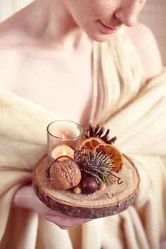 a woman holding a candle and some fruit on a wooden tray with pine cones, an orange slice and cinnamons