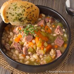 a bowl of soup with ham, beans and bread on a wooden table next to a spoon