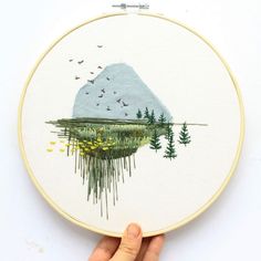 someone is holding up a cross - stitch embroidery project with mountains and trees on it