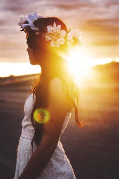 a woman with flowers in her hair is standing on the beach at sundown, looking off into the distance